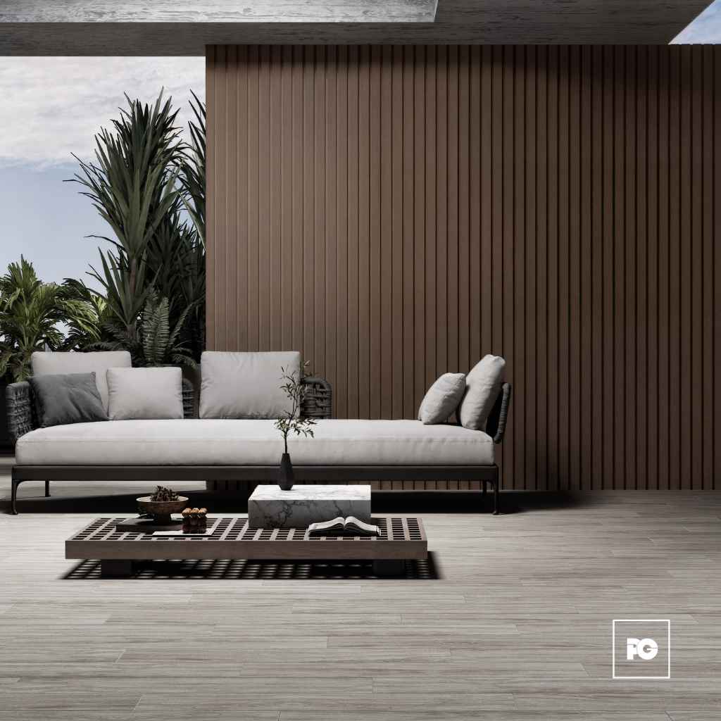 Modern outdoor seating area with a white cushioned sofa, gray pillows, and a low wooden coffee table on a slatted deck, backed by a tall brown slat wall and lush plants to the left.